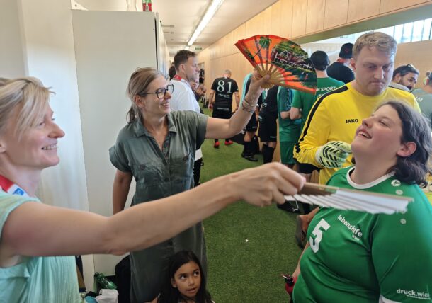 Zwei Frauen fächeln einer lächelnden Frau in einem grünen Sporttrikot Luft zu. In einem hellen Raum mit Grasboden sind weitere Personen zu sehen, darunter Spieler in Uniform und ein Kind.