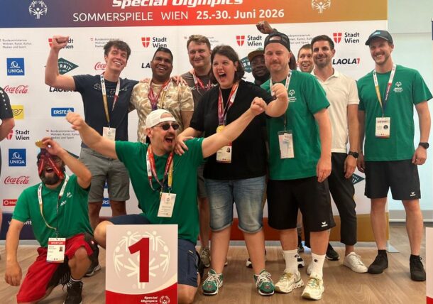 Eine fröhliche Gruppe von Special-Olympics-Athleten und -Trainern feiert gemeinsam in einer Halle und hält vor einem Banner für die Sommerspiele 2026 in Wien ein Schild mit der Aufschrift „1. Platz“ und Medaillen in die Höhe.