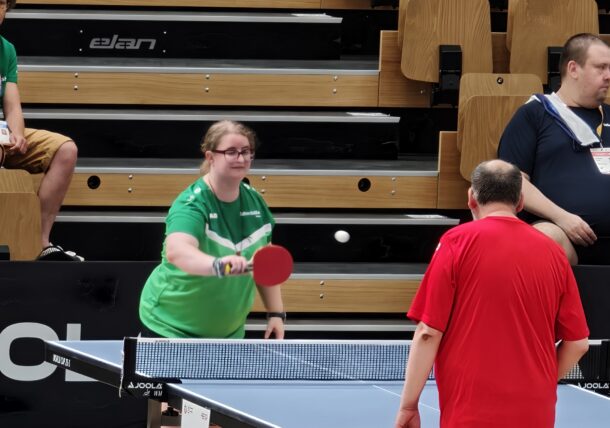 Eine Frau in einem grünen Shirt spielt Tischtennis und schlägt den Ball mit einem roten Schläger, während ihr gegenüber ein Mann in einem roten Shirt steht. Im Hintergrund sind leere Holztribünen und einige sitzende Zuschauer zu sehen.