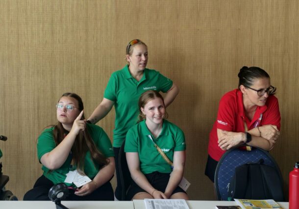 Sechs Personen sitzen oder stehen hinter einem Tisch; drei tragen grüne T-Shirts, drei rote Special-Olympics-T-Shirts. Einige lächeln, eine schaut weg. Auf dem Tisch liegen Papiere und eine rote Wasserflasche. Im Hintergrund ist eine hellbraune Wand zu sehen.