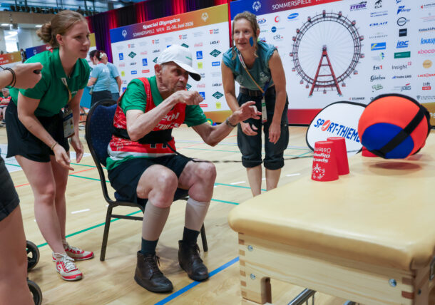 Ein älterer Mann in Sportkleidung wirft, unterstützt von zwei Frauen, bei einer Hallensportveranstaltung vor dem Hintergrund eines Riesenrads einen Basketball auf einen Tisch.