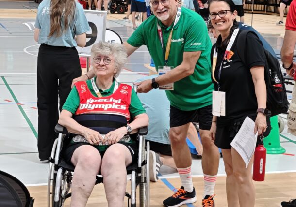 Eine Frau im Rollstuhl und zwei Personen, die neben ihr stehen, lächeln bei einer Hallensportveranstaltung. Im Hintergrund sitzt das Publikum, und auf dem Spielfeld sind mehrere Teilnehmer zu sehen.
