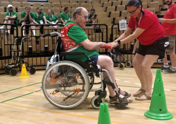 Eine Frau hilft einem Mann im Rollstuhl dabei, in einer Sporthalle zwischen grünen Kegeln hindurchzufahren. Im Hintergrund beobachten Menschen in grünen T-Shirts das Geschehen von den Tribünen aus.