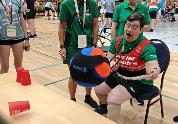 Ein im Rollstuhl sitzender Sportler mit Down-Syndrom fängt während einer Special-Olympics-Hallenveranstaltung begeistert einen großen, bunten Ball, während Teamkollegen und Betreuer in der Nähe stehen. Auf dem Tisch vor ihnen stehen rote Becher.