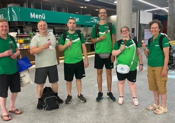 Sechs Personen stehen in einem Innenraum neben Einkaufswagen, lächeln und halten Eiswaffeln in der Hand. Sie tragen grüne Oberteile und legere Hosen. Hinter ihnen sind eine Speisekarte und eine Snackbar zu sehen. Der Schauplatz scheint ein Flughafen oder eine Cafeteria zu sein.