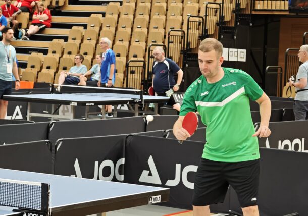 Ein Mann in einem grünen Hemd macht sich bereit, einen Tischtennisball zu schlagen, während im Hintergrund Zuschauer in einer Halle zuschauen.