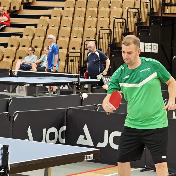 Ein Mann in einem grünen Hemd macht sich bereit, einen Tischtennisball zu schlagen, während im Hintergrund Zuschauer in einer Halle zuschauen.