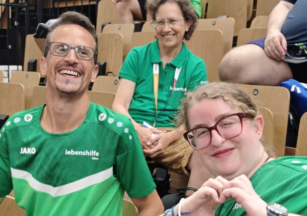 Drei Personen in grünen T-Shirts sitzen in einem Saal. Die beiden vorne lächeln, wobei eine von ihnen mit den Händen ein Herz formt. Hinter ihnen sitzt eine weitere Person in Grün, die ebenfalls lächelt. Die Atmosphäre ist fröhlich und freundlich.