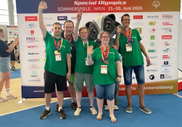 Fünf Personen in grünen T-Shirts mit Medaillen lächeln und winken vor einem Banner der Special Olympics Wien 2026 in einer Sporthalle. Im Hintergrund sind ein Maskottchen und die Logos der Sponsoren zu sehen.