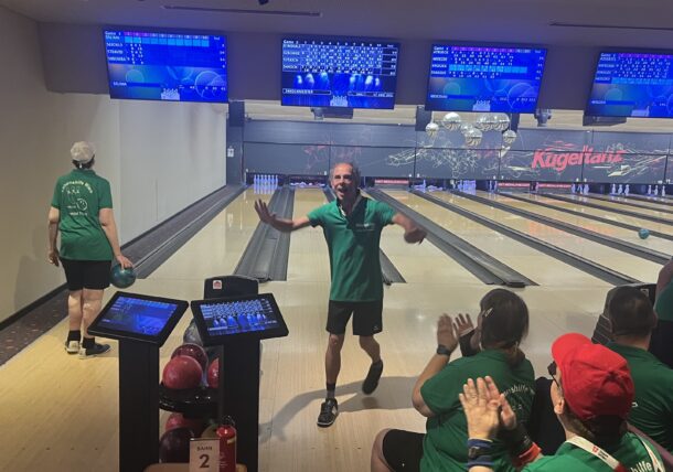 Ein Mann jubelt beim Bowling, während seine Teamkollegen in grünen Trikots zusehen und ihn anfeuern. Auf der Bahn daneben spielt eine weitere Person. Die Bowling-Ergebnisse werden auf Monitoren über den Bahnen angezeigt.