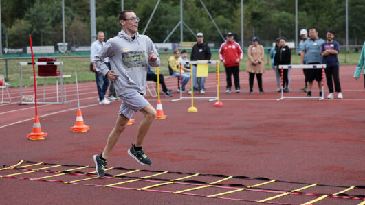 Ein Mann in Sportkleidung läuft lächelnd über eine Agility-Leiter auf einer Laufbahn. Mehrere Zuschauer stehen am Rand und feuern ihn an. Orangefarbene Kegel markieren den Parcours.