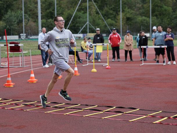 Ein Mann in Sportkleidung läuft lächelnd über eine Agility-Leiter auf einer Laufbahn. Mehrere Zuschauer stehen am Rand und feuern ihn an. Orangefarbene Kegel markieren den Parcours.