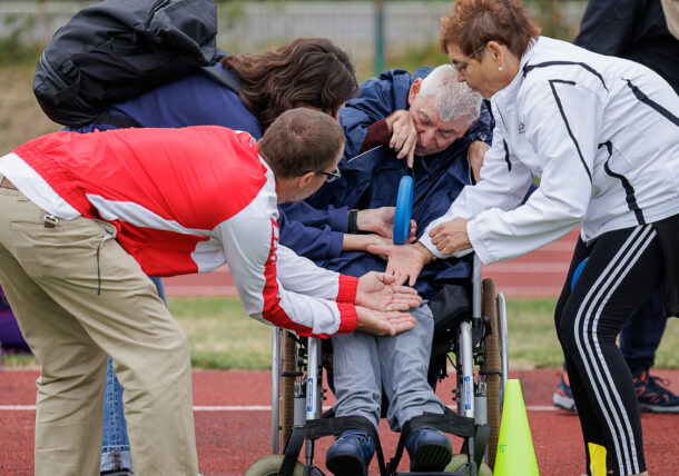 Eine Gruppe von Menschen unterstützt und feuert einen älteren Mann im Rollstuhl an, der an einer Sportveranstaltung im Freien auf einer Laufbahn teilnimmt, die von Kegeln und Rasen umgeben ist.