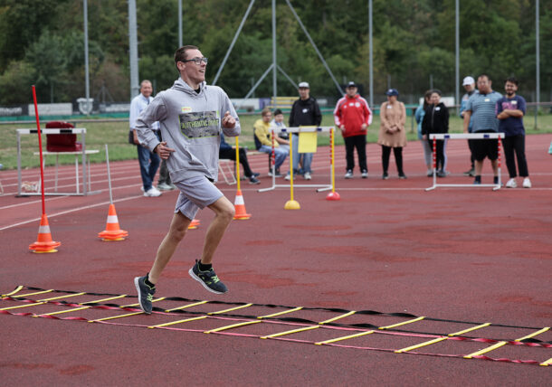 Ein Mann in Sportkleidung läuft lächelnd über eine Agility-Leiter auf einer Laufbahn. Im Hintergrund schauen mehrere Menschen zu und feuern ihn an. Rund um den Bereich sind orangefarbene Kegel und Absperrungen aufgestellt.