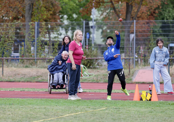 Ein Junge wirft auf einem Leichtathletikplatz einen Ball, während eine Frau daneben steht. Im Hintergrund lächeln zwei Personen, von denen eine im Rollstuhl sitzt. Eine weitere Person steht seitlich daneben. Orangefarbene Kegel markieren den Platz.