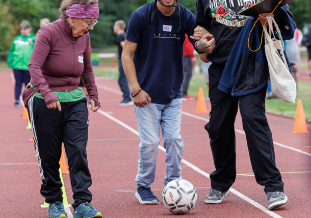 Drei Menschen lächeln auf einer Laufbahn; einer von ihnen trägt einen Helm und kickt sanft gegen einen Fußball. Die anderen schauen zu und feuern ihn an, umgeben von orangefarbenen Kegeln und Grünflächen im Hintergrund.