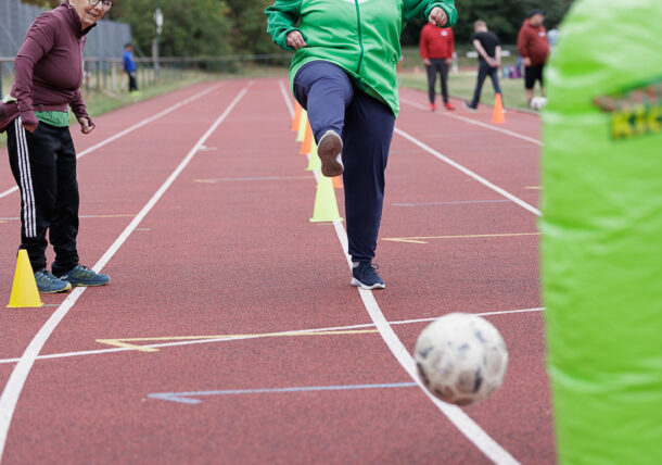 Eine Person in einer grünen Jacke kickt einen Fußball auf einer Laufbahn, während eine andere Person daneben steht. Entlang der Laufbahn sind gelbe Kegel und grüne gepolsterte Absperrungen aufgestellt. Im Hintergrund sind Bäume zu sehen.