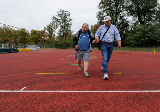 Zwei Männer gehen Hand in Hand nebeneinander auf einer roten Laufbahn im Freien. Auf der Laufbahn sind mehrere gelbe und rote Streifen zu sehen. Im Hintergrund sind Bäume und Sportgeräte zu erkennen.