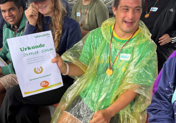 Ein lächelnder Mann in einem gelbgrünen Poncho hält eine Urkunde in der Hand, während er zusammen mit anderen bei einer Veranstaltung im Freien sitzt. Einige tragen Medaillen, und alle wirken fröhlich und begeistert.