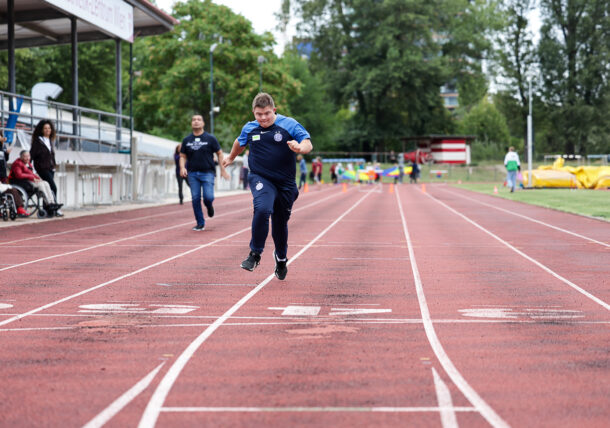 Ein junger Sportler in blauer Sportbekleidung sprintet auf einer Laufbahn und nähert sich der Ziellinie. Im Hintergrund sind weitere Teilnehmer und Zuschauer zu sehen.