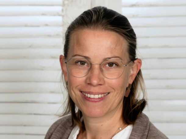 Eine lächelnde Frau mit einer Brille mit dünnem Rahmen vor einem weißen Lamellenhintergrund.