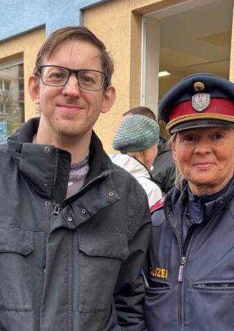 Ein Mann mit Brille und schwarzer Jacke, der neben einem uniformierten österreichischen Polizisten steht.