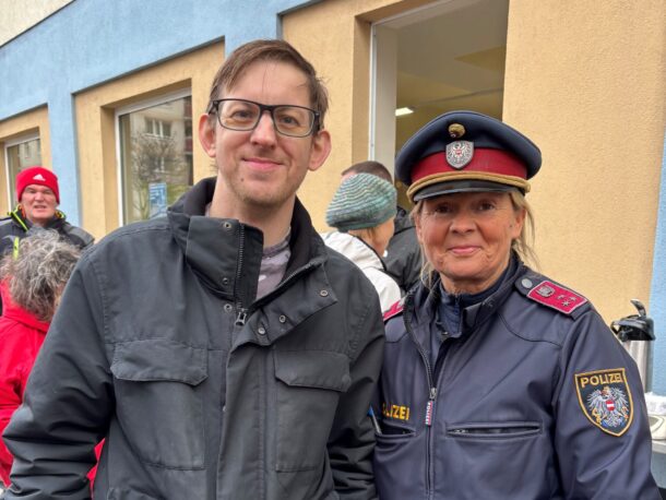 Ein Mann mit Brille und schwarzer Jacke, der neben einem uniformierten österreichischen Polizisten steht.