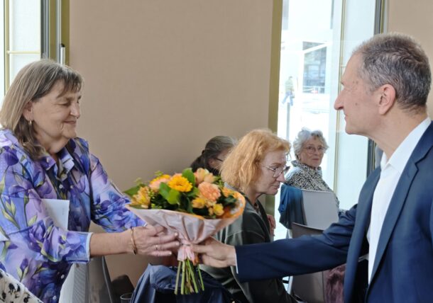 Ein Mann im Anzug überreicht einer lächelnden Frau in einer geblümten Bluse einen Strauß aus orangefarbenen und gelben Blumen, während im Hintergrund andere Personen sitzen.