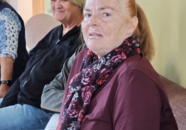 Zwei Frauen sitzen nebeneinander; die Frau im Vordergrund trägt ein weinrotes Langarmhemd und einen schwarz-roten Schal mit Blumenmuster.
