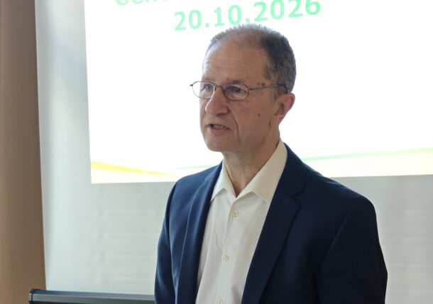 Ein Mann in Anzugjacke und weißem Hemd steht vor einer Präsentationsleinwand mit der Aufschrift Generalversammlung 20.10.2026 und hält ein Blatt Papier in der Hand.