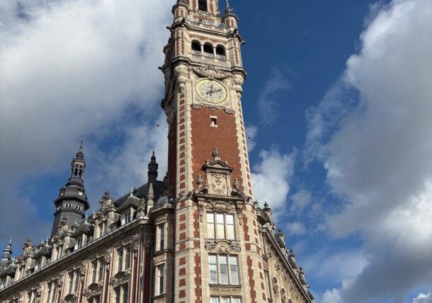 Der kunstvoll verzierte Backstein-Glockenturm der Handelskammer in Lille, Frankreich, von unten gesehen vor einem blauen Himmel mit Wolken, im Vordergrund rote Regenschirme.
