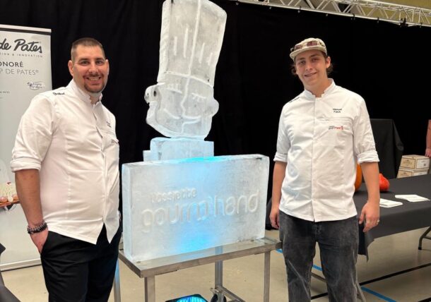 Zwei Köche stehen zu beiden Seiten einer Eisskulptur in Form einer Kochmütze, die auf einem Sockel steht, auf dem die Aufschrift Rossette gourmand zu lesen ist.