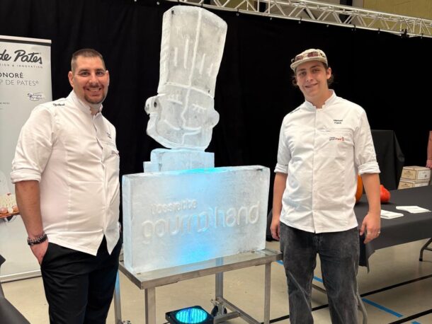 Zwei Köche stehen zu beiden Seiten einer Eisskulptur in Form einer Kochmütze, die auf einem Sockel steht, auf dem die Aufschrift Rossette gourmand zu lesen ist.