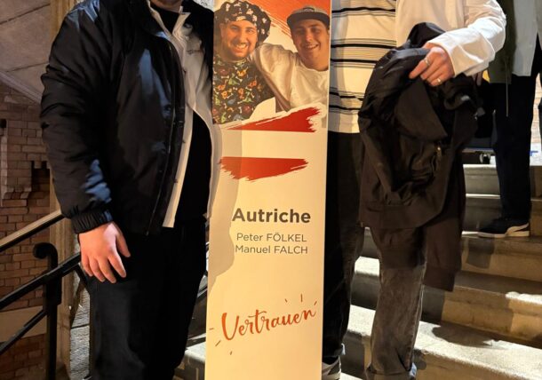 Zwei Männer halten ein hohes, senkrechtes Transparent hoch, auf dem ein Foto von zwei Personen, der Text Autriche, Peter FÖLKEL, Manuel FALCH, Vertrauen sowie das Logo von Lassiette Gourmande zu sehen sind.
