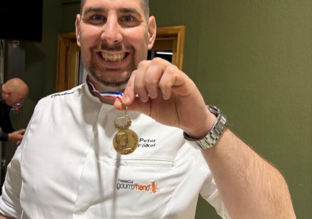 Ein lächelnder Koch in weißer Kochuniform hält eine Goldmedaille hoch.