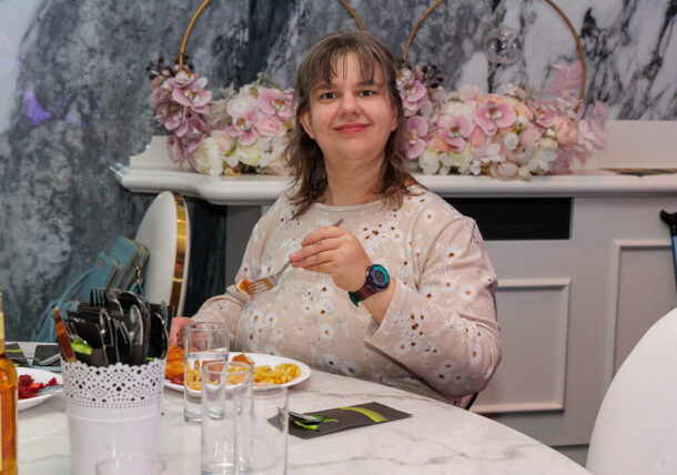Eine Frau mit schulterlangem Haar sitzt an einem Tisch, hält eine Gabel in der Hand und isst Pommes und Fleisch.