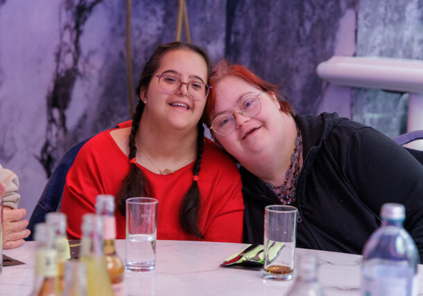 Zwei Frauen mit Down-Syndrom lächeln, während sie ihre Köpfe an einem Tisch aneinanderlehnen.