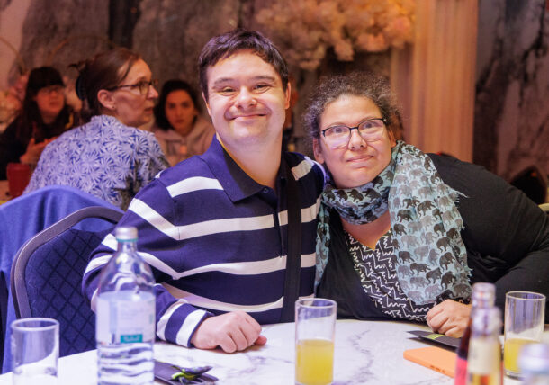 Ein Mann in einem gestreiften Hemd und eine Frau mit Brille und einem gemusterten Schal sitzen an einem Tisch und lächeln einander an.