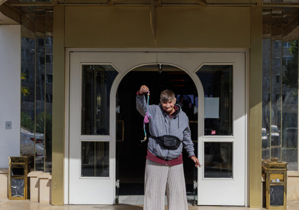 Eine Person in einem grauen Kapuzenpulli und einer Hose mit weitem Bein steht im Eingangsbereich eines Hotels mit der Aufschrift PORTOFINO und hält einen kleinen bunten Schlüsselanhänger hoch.