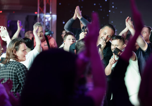 Ein Mann singt bei einer Veranstaltung in einer Halle in ein Mikrofon, während er von einer Menschenmenge umringt ist; einige Zuschauer jubeln, andere nehmen das Geschehen mit ihren Handys auf.