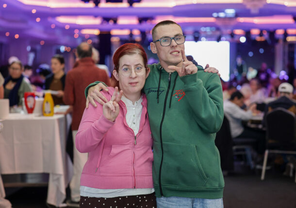 Eine junge Frau in einem rosa Kapuzenpulli und ein junger Mann in einem grünen Kapuzenpulli posieren gemeinsam bei einer Veranstaltung und machen dabei beide Handgesten.