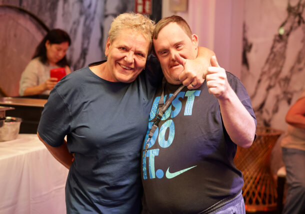 Eine lächelnde Frau mit kurzen blonden Haaren und ein Mann in einem Just Do It-T-Shirt umarmen sich, während sie für ein Foto posieren.