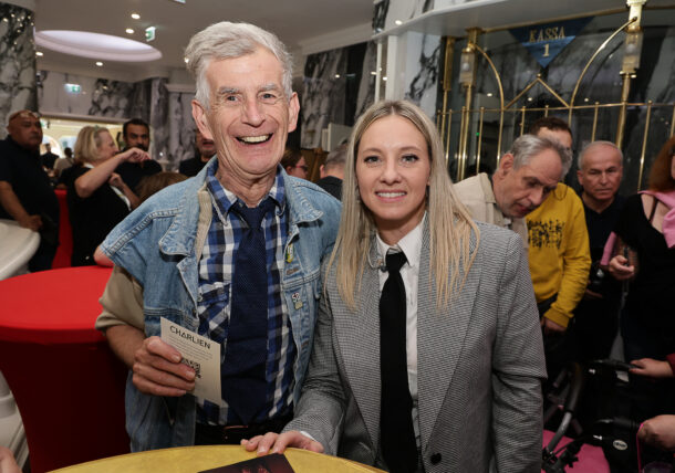 Ein älterer Mann in einer Jeansweste und eine jüngere Frau in einem Blazer stehen bei einer Veranstaltung nebeneinander und lächeln.